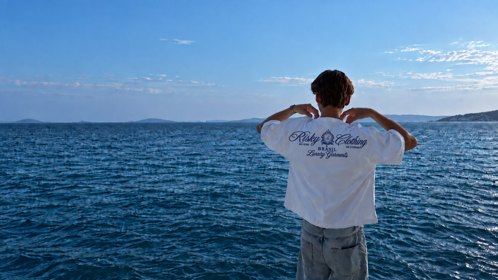 Rapaz de costas olhando o mar, vestindo a camiseta branca Coat of Arms, com o brasão Risky Clothing azul marinho nas costas