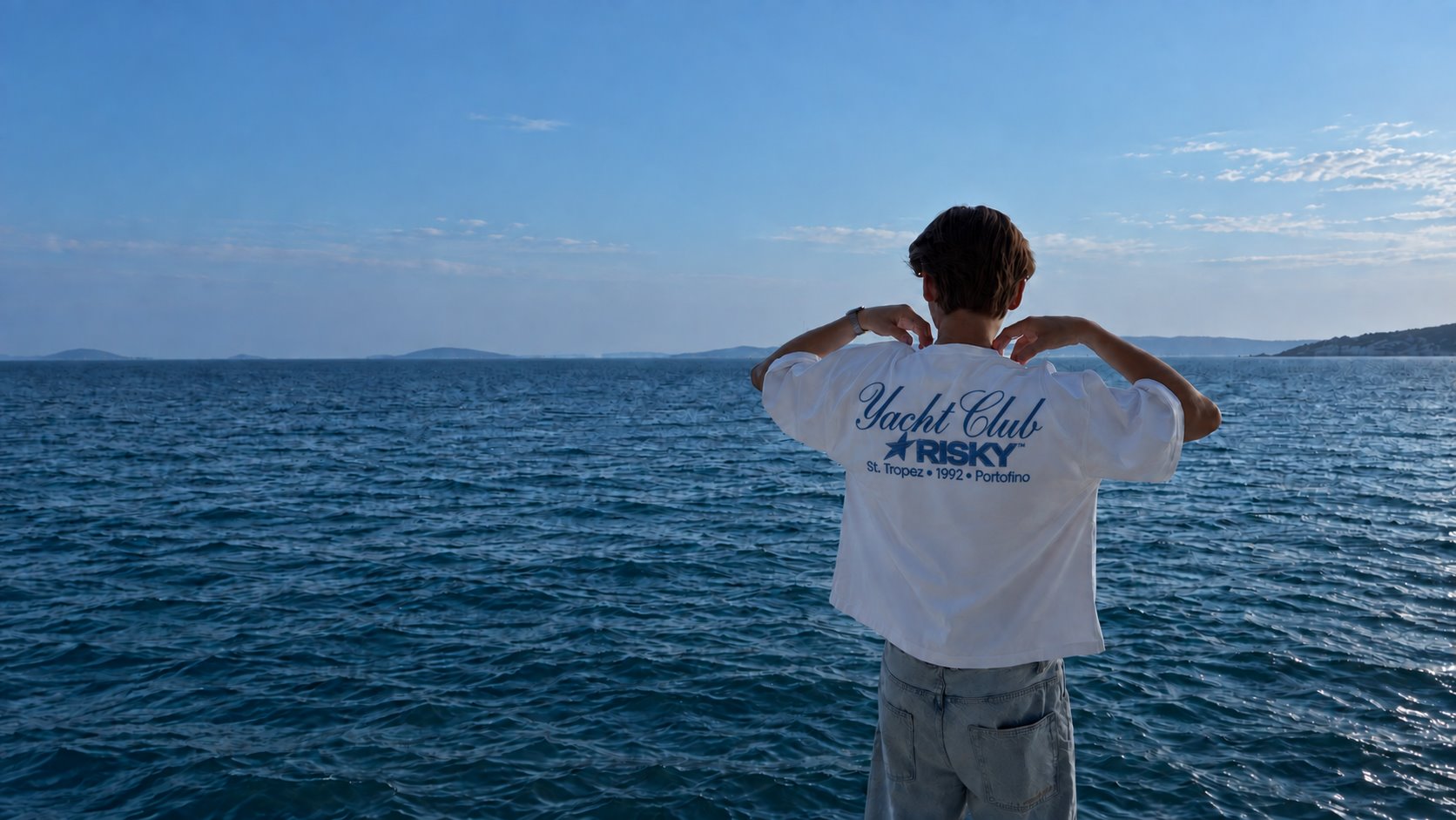 Rapaz de costas olhando o mar, vestindo a camiseta branca Yacht Club, com a estrela azul estampada nas costas