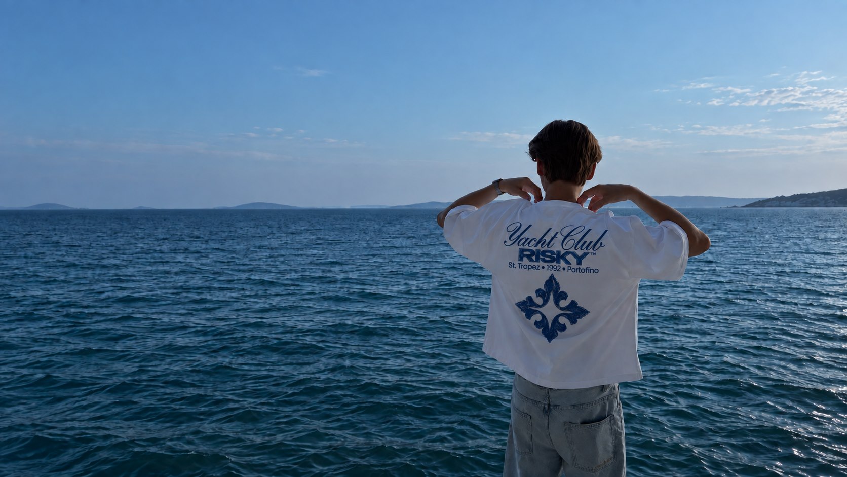 Rapaz de costas olhando o mar, vestindo a camiseta branca Yacht Club, com a estrela azul estampada nas costas