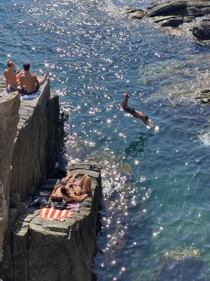 Pessoas tomando sol nas rochas enquanto um rapaz mergulha no mar brilhante