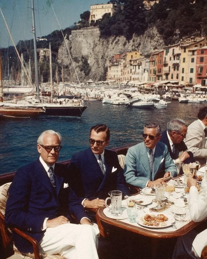 Foto antiga de homens de terno almoçando num barco no porto de Portofino, com as casas coloridas ao fundo